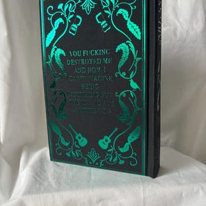 Puede incluir: Un libro de tapa dura negro con detalles en l&aacute;mina de color turquesa. La portada presenta dise&ntilde;os florales, guitarras y el texto "YOU FUCKING DESTROYED ME AND NOW I CAN'T IMAGINE BEING ANYTHING BUT THE MAN I AM WITH YOU."