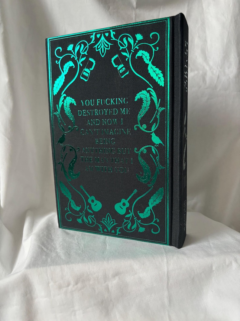 Puede incluir: Un libro de tapa dura negro con detalles en l&aacute;mina de color turquesa. La portada presenta dise&ntilde;os florales, guitarras y el texto "YOU FUCKING DESTROYED ME AND NOW I CAN'T IMAGINE BEING ANYTHING BUT THE MAN I AM WITH YOU."