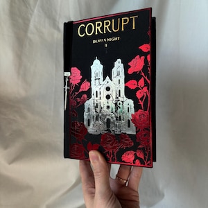 May include: A black book with the title "CORRUPT" in gold lettering and a silver illustration of a building. Red roses and foliage decorate the cover. The book is held by a person.