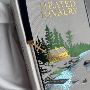 May include: A hardcover book titled "HEATED RIVALRY" with a forest and lake scene. The book has a navy blue velvet bookmark with a gold-colored cross charm. The cover is light gray with gold lettering.
