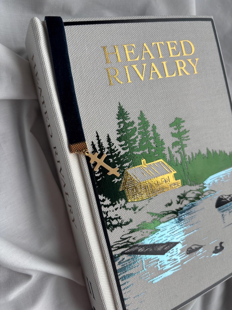 May include: A hardcover book titled "HEATED RIVALRY" with a forest and lake scene. The book has a navy blue velvet bookmark with a gold-colored cross charm. The cover is light gray with gold lettering.