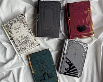 Könnte beinhalten: Fünf Hardcover-Bücher mit dekorativen Einbänden liegen auf einer weißen Oberfläche. Die Bücher haben goldfarbene Kanten und enthalten Titel wie "A Court of Thorns and Roses". Jedes Buch hat ein Leseband.