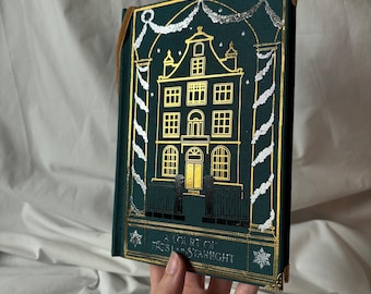 Könnte beinhalten: Ein Hardcover-Buch mit tealfarbenem Einband und einer gold- und silberfarbenen Illustration eines Gebäudes. Der Titel "A Court of Frost and Starlight" ist unten aufgedruckt. Das Buch hat goldfarbene Kanten und ein braunes Lesezeichen.