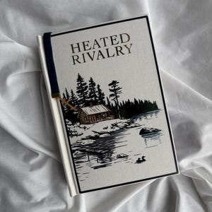 May include: A hardcover book titled "HEATED RIVALRY" with a landscape illustration. The cover features a cabin, trees, and a lake scene in black, white, and green. A navy blue bookmark with a gold charm is attached.