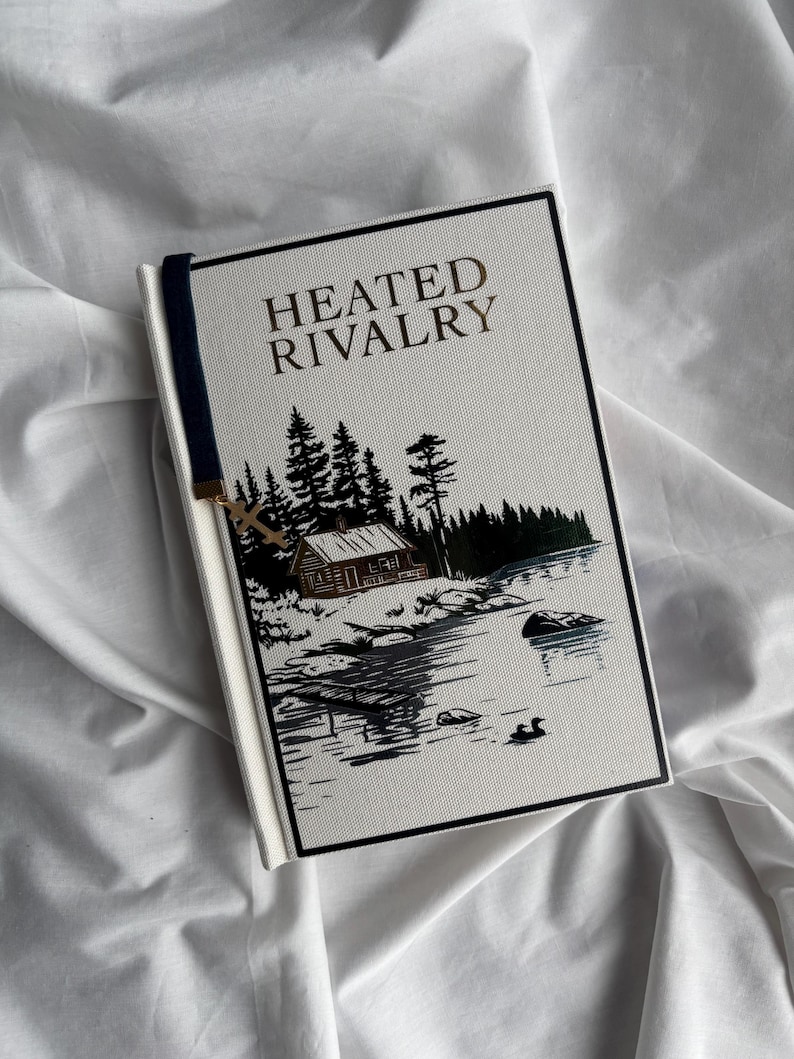 May include: A hardcover book titled "HEATED RIVALRY" with a landscape illustration. The cover features a cabin, trees, and a lake scene in black, white, and green. A navy blue bookmark with a gold charm is attached.