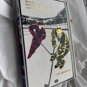 May include: A hardcover journal with a hockey theme. The cover features two hockey players in red and gold, with the text "does it feel like agony for you too?" and "NOT ANYMORE" in gold lettering. The journal's spine has a blue and white pattern.