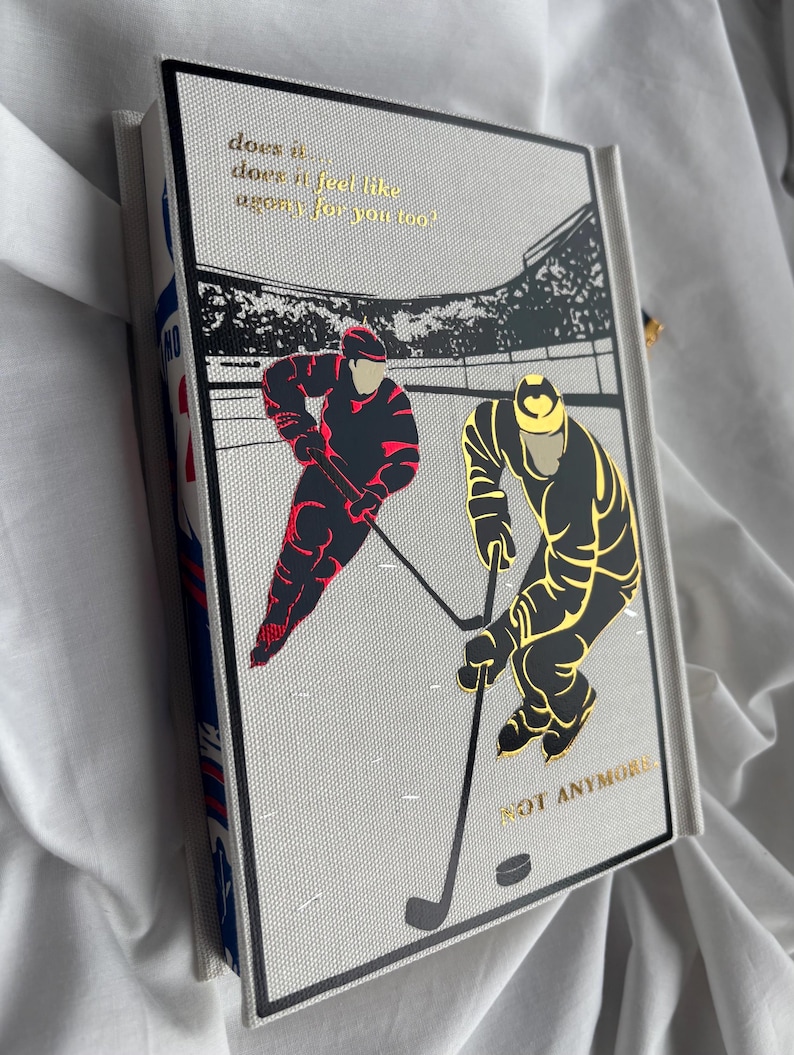May include: A hardcover journal with a hockey theme. The cover features two hockey players in red and gold, with the text "does it feel like agony for you too?" and "NOT ANYMORE" in gold lettering. The journal's spine has a blue and white pattern.