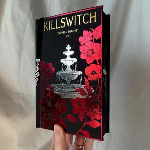 Puede incluir: Un libro negro con el título "KILLSWITCH" en letras doradas. La portada presenta una ilustración de fuente plateada y detalles florales rojos. El libro es parte de la serie "Devils Night III".