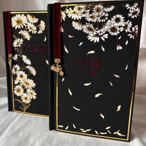 May include: Two hardback books with black covers, gold edging, and floral patterns. The covers display white and gold daisies, with the title "TORMENT" in red. Each book has a claret ribbon bookmark with a gold charm.