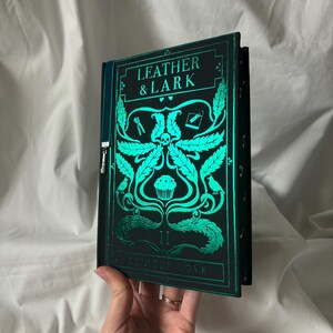 Puede incluir: Un libro verde oscuro con el texto "LEATHER & LARK" e ilustraciones turquesas. La portada presenta una calavera, hojas y un dise&ntilde;o de cupcake. El t&iacute;tulo "RUNIC LOVE" tambi&eacute;n es visible.