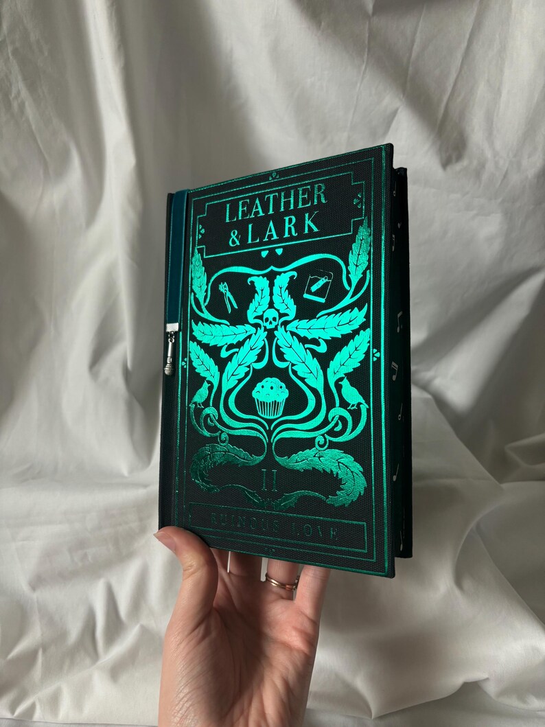 Puede incluir: Un libro verde oscuro con el texto "LEATHER & LARK" e ilustraciones turquesas. La portada presenta una calavera, hojas y un dise&ntilde;o de cupcake. El t&iacute;tulo "RUNIC LOVE" tambi&eacute;n es visible.
