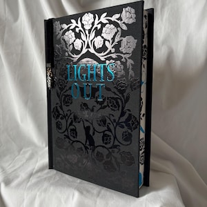 Può includere: Un libro con copertina rigida nera con un motivo floreale e le parole "LIGHTS OUT" in turchese. Il libro presenta un motivo floreale argentato e un dorso nero. Un ciondolo è appeso al libro.