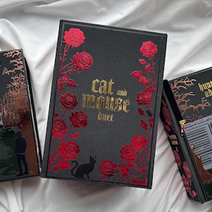 May include: Three black books with metallic accents. The left and right books are titled Haunting Adeline and feature illustrations of a house and a train car. The center book is titled Cat and Mouse Duet and features red roses and a black cat.