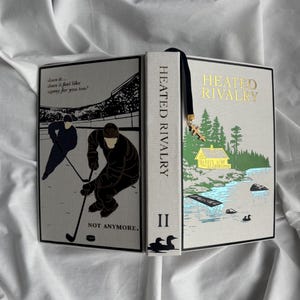 May include: A hardcover book titled "Heated Rivalry II" with a black bookmark and a gold charm. The cover features illustrations of hockey players and a lakeside scene with a yellow cabin. The spine reads "Heated Rivalry".