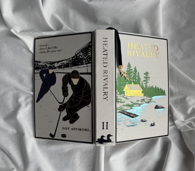 May include: A hardcover book titled "Heated Rivalry II" with a black bookmark and a gold charm. The cover features illustrations of hockey players and a lakeside scene with a yellow cabin. The spine reads "Heated Rivalry".