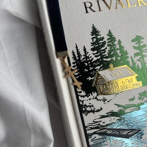 May include: A black velvet bookmark with a gold cross charm is placed in a book. The book cover is light gray with gold lettering and a forest scene illustration in green and gold.