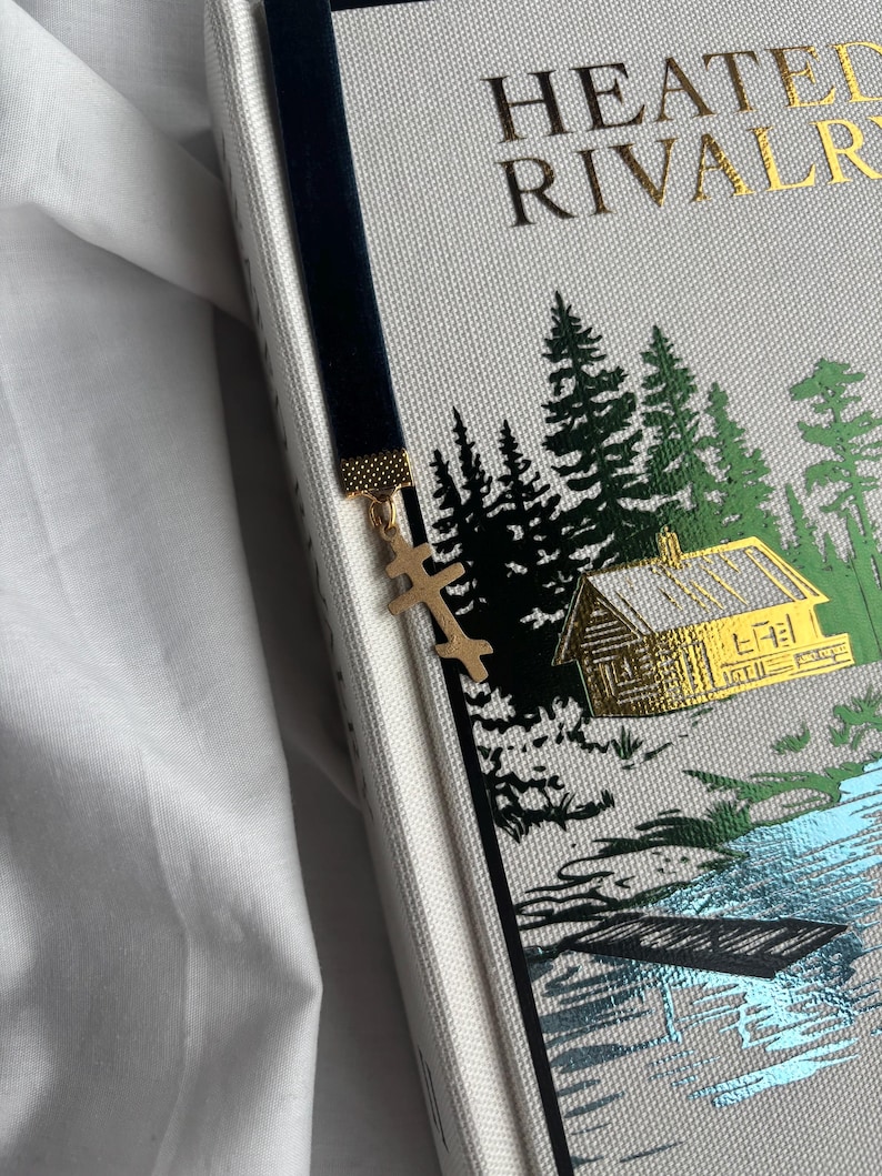 May include: A black velvet bookmark with a gold cross charm is placed in a book. The book cover is light gray with gold lettering and a forest scene illustration in green and gold.
