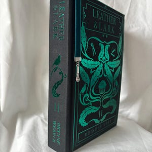 Puede incluir: Un libro de tapa dura titulado "Leather & Lark: Ruinous Love II" con letras e ilustraciones verde azulado sobre un fondo negro. El lomo presenta el t&iacute;tulo y el nombre del autor. Se inserta un marcap&aacute;ginas verde azulado con un dije plateado.
