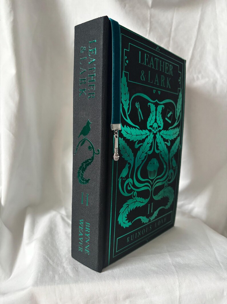 Puede incluir: Un libro de tapa dura titulado "Leather & Lark: Ruinous Love II" con letras e ilustraciones verde azulado sobre un fondo negro. El lomo presenta el t&iacute;tulo y el nombre del autor. Se inserta un marcap&aacute;ginas verde azulado con un dije plateado.