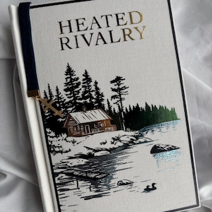 May include: A hardcover book titled "HEATED RIVALRY" with gold lettering. The cover features a black and white illustration of a cabin by a lake with trees. A navy blue bookmark with a gold cross charm is attached.