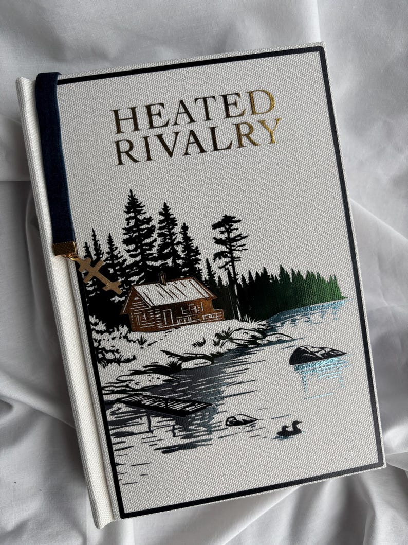 May include: A hardcover book titled "HEATED RIVALRY" with gold lettering. The cover features a black and white illustration of a cabin by a lake with trees. A navy blue bookmark with a gold cross charm is attached.
