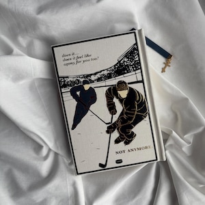 May include: A hardcover book with a black and gold illustration of two hockey players on a white background. The text on the cover reads "does it... does it feel like agony for you too?" and "NOT ANYMORE". A blue bookmark with a gold cross is attached.