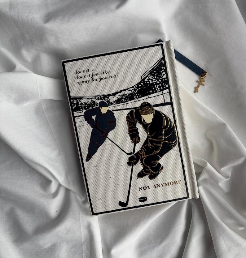 May include: A hardcover book with a black and gold illustration of two hockey players on a white background. The text on the cover reads "does it... does it feel like agony for you too?" and "NOT ANYMORE". A blue bookmark with a gold cross is attached.