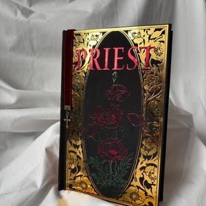 May include: A black book with a gold and red embossed design. The title "PRIEST" is written in red lettering. A red velvet bookmark with a silver cross charm is attached to the book.