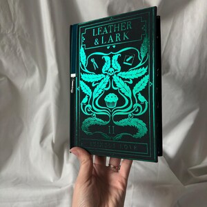 Puede incluir: Un libro de tapa dura titulado "LEATHER & LARK" con una cubierta verde azulado y negra. La cubierta presenta ilustraciones verde azulado de una calavera, hojas y otros elementos decorativos. El libro est&aacute; sostenido en una mano.