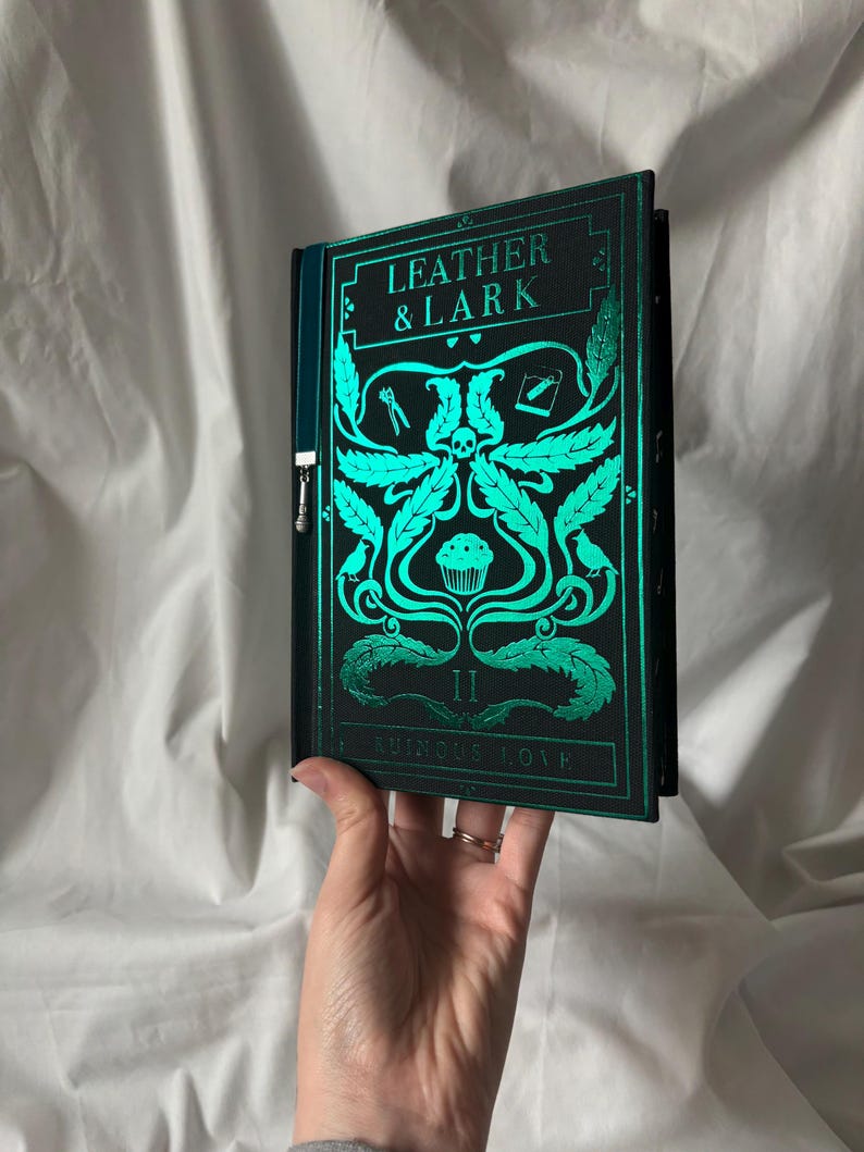 Puede incluir: Un libro de tapa dura titulado "LEATHER & LARK" con una cubierta verde azulado y negra. La cubierta presenta ilustraciones verde azulado de una calavera, hojas y otros elementos decorativos. El libro est&aacute; sostenido en una mano.