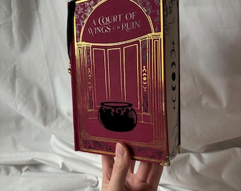 Könnte beinhalten: Ein weinrotes Buch mit goldenen Akzenten und dem Titel "A Court of Wings and Ruin". Das Cover zeigt ein goldenes Bogendesign mit einem schwarzen Kessel. Der Buchrücken hat ein schwarzes Leseband.