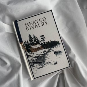 May include: A hardcover book titled "HEATED RIVALRY" with a landscape illustration of a cabin, trees, and a lake. The book has a navy blue bookmark and is set against a white fabric background.