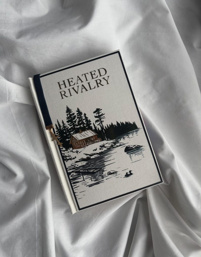 May include: A hardcover book titled "HEATED RIVALRY" with a landscape illustration of a cabin, trees, and a lake. The book has a navy blue bookmark and is set against a white fabric background.
