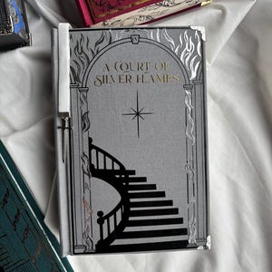 Puede incluir: Un libro titulado "A Court of Silver Flames" con una cubierta gris que presenta un diseño de escalera negra. El título está en letras doradas. El libro tiene un marcapáginas de cinta blanca y protectores de esquina plateados. Otros libros son visibles en el fondo.