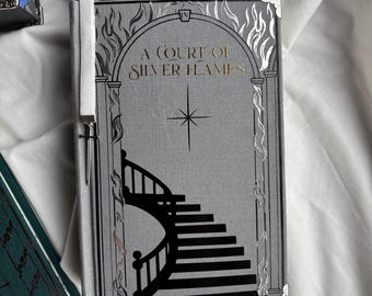 Könnte beinhalten: Ein Buch mit dem Titel "A Court of Silver Flames" mit einem grauen Einband mit einem schwarzen Treppendesign. Der Titel ist in goldener Schrift. Das Buch hat ein weißes Band-Lesezeichen und silberne Eckenschützer. Andere Bücher sind im Hintergrund sichtbar.
