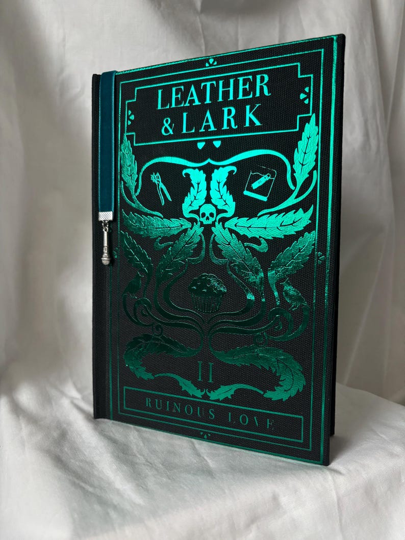 Puede incluir: Un libro de tapa dura titulado "LEATHER & LARK II RUINOUS LOVE" con una cubierta negra y un dise&ntilde;o en relieve verde azulado. El dise&ntilde;o incluye una calavera, hojas y herramientas. Una cinta marcap&aacute;ginas verde azulado con un dije plateado est&aacute; adjunta.