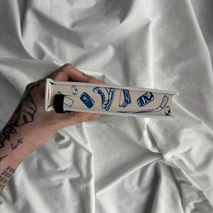 May include: A rectangular, off-white box with a blue illustration of sports equipment, including a hockey skate, a can, and a hockey puck. The box has a black strap and a canvas-like texture.