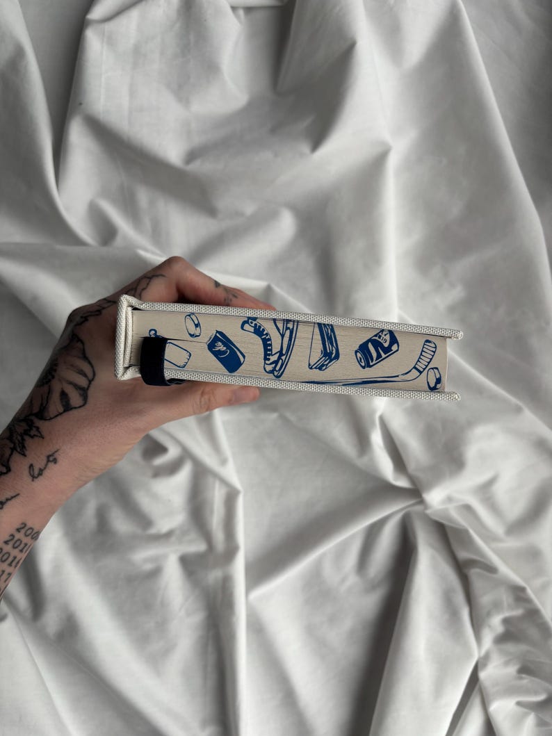 May include: A rectangular, off-white box with a blue illustration of sports equipment, including a hockey skate, a can, and a hockey puck. The box has a black strap and a canvas-like texture.