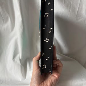 Puede incluir: Lomo de libro negro decorado con notas musicales blancas. Se ve un marcap&aacute;ginas de cinta azul. El libro se sostiene en una mano sobre un fondo blanco.