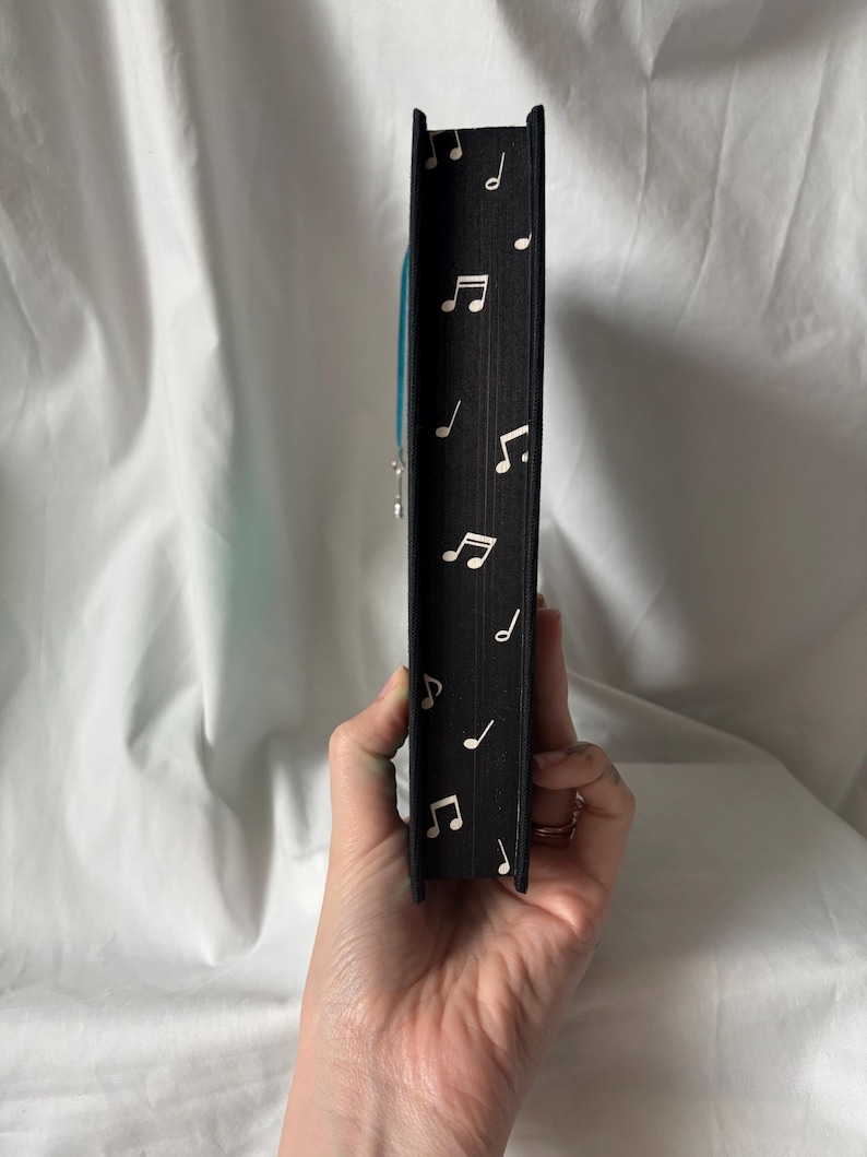 Puede incluir: Lomo de libro negro decorado con notas musicales blancas. Se ve un marcap&aacute;ginas de cinta azul. El libro se sostiene en una mano sobre un fondo blanco.