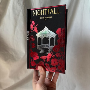 May include: A black hardcover book titled "NIGHTFALL" with "DEVIL'S NIGHT IV" printed below. The cover features a silver gazebo illustration and red floral accents. A silver bookmark is attached.