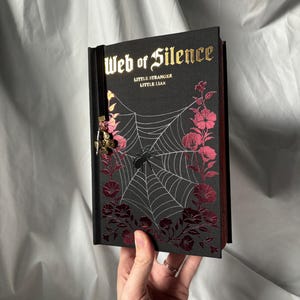 May include: A black book titled "Web of Silence" with gold lettering and a spiderweb design. Pink floral accents frame the web. A black ribbon bookmark with a charm is attached.