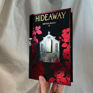 May include: A black book with the title "HIDEAWAY" in gold lettering. The cover features a silver and red floral design with the text "DEVILS NIGHT II". The book is held open, revealing the inside pages.