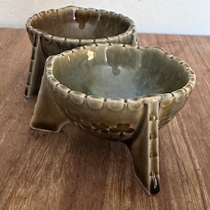 May include: Two green ceramic bowls with a scalloped rim and three legs each. The bowls have a floral design and are sitting on a wooden surface.