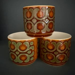 May include: Three vintage ceramic cups with a repeating pattern of olive green and orange shapes on a brown background. The cups are stacked, showcasing their design and colour variations. The interior of the cups is a cream colour.