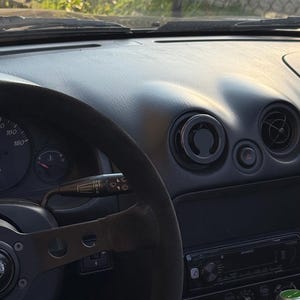 May include: Car interior showing a black steering wheel with a suede grip and a Momo logo. The dark grey dashboard includes air vents, gauges, and a Kenwood stereo. The image is taken in daylight, with a view of the windscreen.