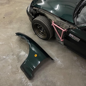 May include: A dark green sports car with a missing fender and a pink support structure. The car has a black tire and a gold-colored wheel. The fender is detached and lying on the ground. The car has a sticker that says "RACING MASSIVE".