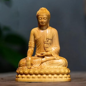 May include: A wooden Buddha statue in a seated meditation pose. The figure is carved from light-coloured wood, with intricate details in the robe, hair, and lotus base. The statue has a serene expression and is a symbol of peace and enlightenment.