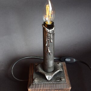 Puede incluir: Una lámpara de vela de metal negro con una base de madera. La lámpara tiene un diseño decorativo de goteo de metal y una bombilla de vidrio transparente.