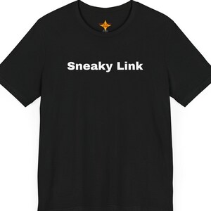 May include: Black t-shirt with the text "Sneaky Link" in white, centered on the chest. The shirt has short sleeves and a crew neck. A small, gold-colored emblem is visible near the neckline.